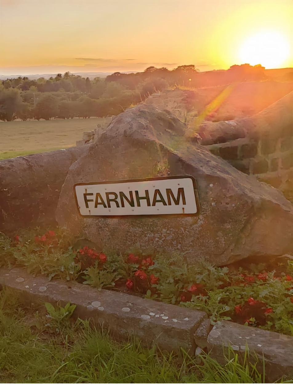The Farnham village welcome stone at sunset — a large rough boulder set into a low limestone wall, with the village name on a small white plate, planted around with red flowers, the sun setting low and golden behind it over the village green