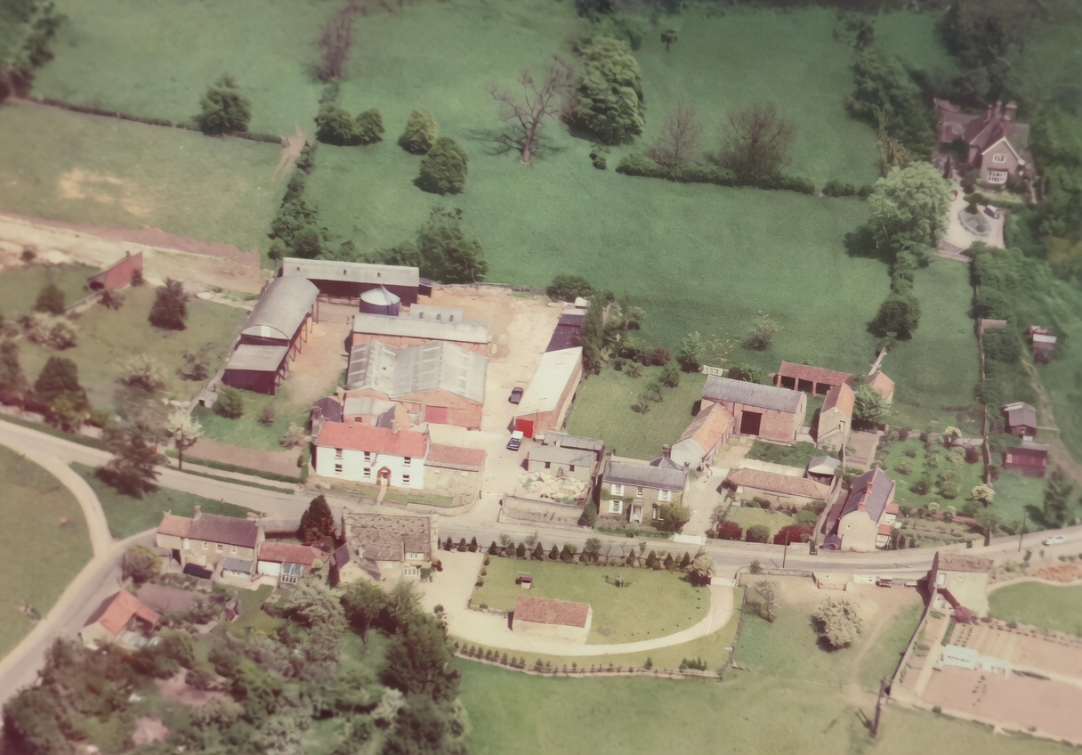 Aerial colour photograph showing Farnham's working farmstead with grain silo and barns at upper left, the cluster of stone houses around the village green, the curving main road, and Manor Farm, Farnham House and Farnham Grange visible to the right. Photographed in the 1960s or early 1970s, before Beech Close was built.