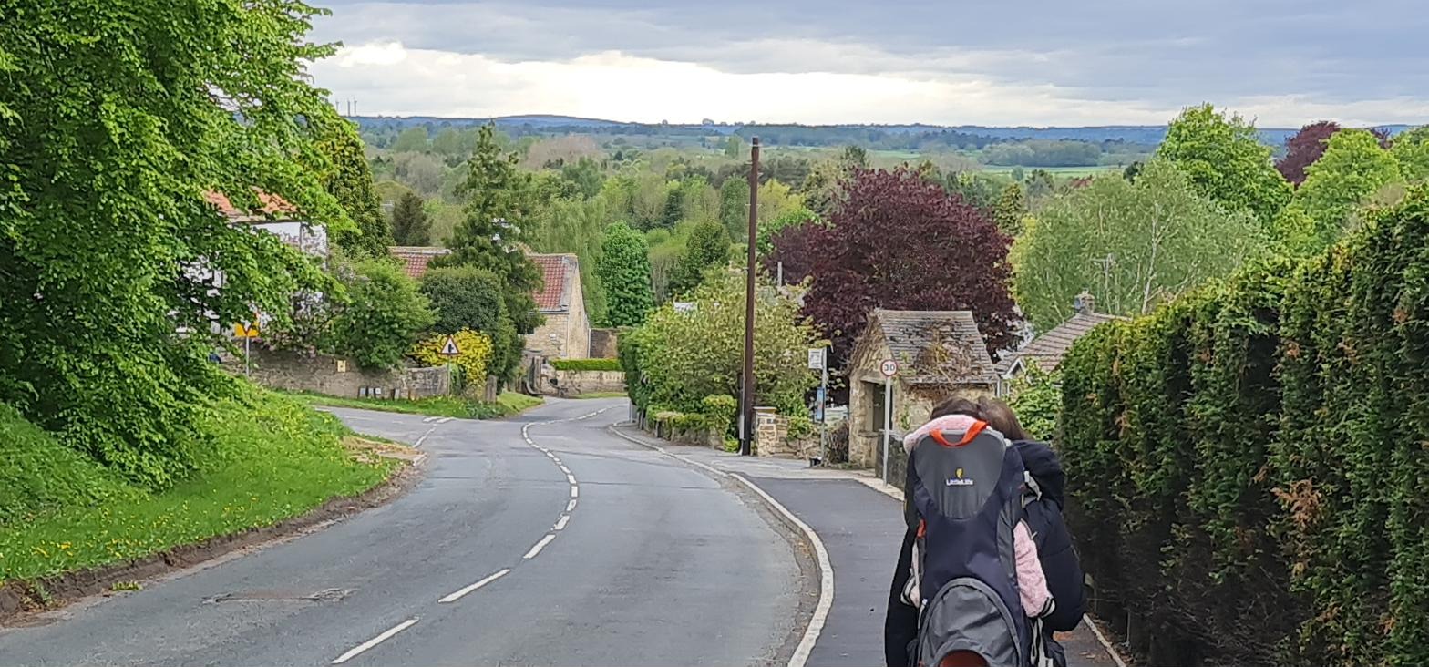 A walker carrying a child in a back-carrier descends Farnham Lane in late spring, with the village rooftops and open countryside visible beyond