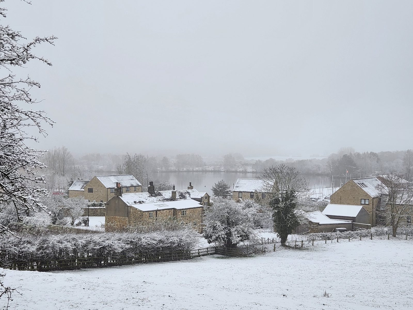 A panoramic snowy winter view from above Farnham — stone houses with snow-covered roofs, frost-laden trees and hedgerows in the middle distance, a snow-covered meadow in the foreground, and the calm lakes and marina just visible in the misty far distance
