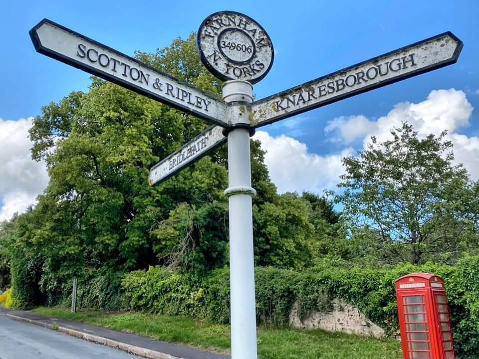 The cast-iron fingerpost at Farnham showing directions to Scotton and Ripley, Knaresborough, and the bridlepath, with a red telephone kiosk behind