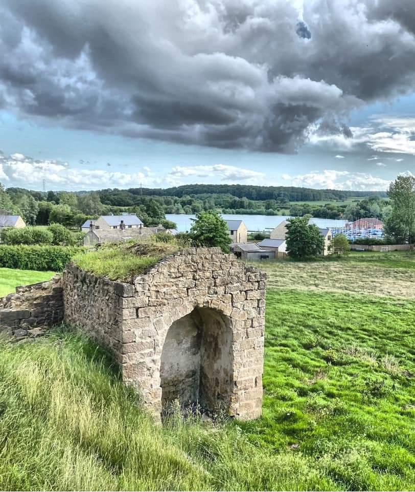 A weathered stone archway in long summer grass with the Farnham lakes, the marina, and the village stone houses visible in the background, under a sky of dramatic cumulus cloud