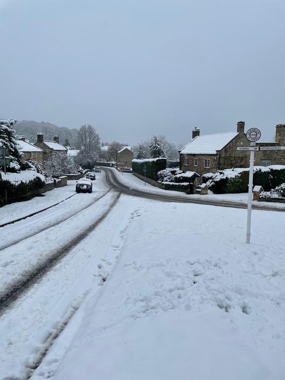Farnham's white-painted Yorkshire fingerpost on a snowy verge, pointing to Knaresborough and Scotton & Ripley, with the village's stone houses to the right and tyre tracks running down the lane