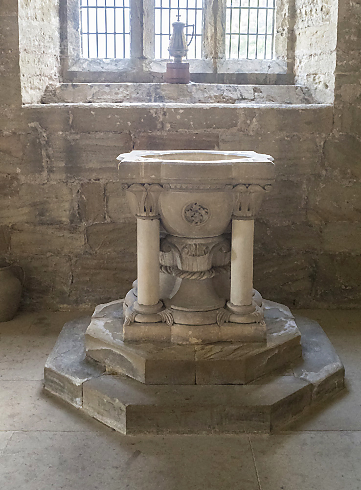 The octagonal Norman font of St Oswald's Church, carved in pale limestone with rosettes and a fluted basin, standing on three columns above a stepped octagonal stone base, with two leaded windows behind