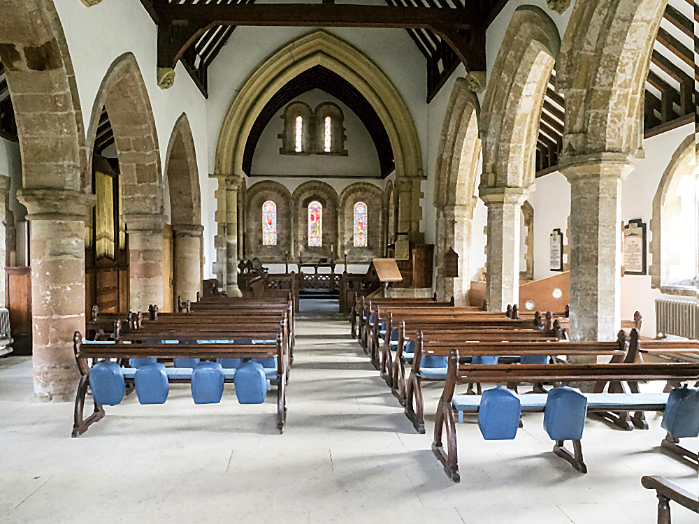 The east end of St Oswald's nave from closer up, the chancel arch framing the three Norman round-headed windows behind, with rows of wooden pews and a stone flagged floor in the foreground