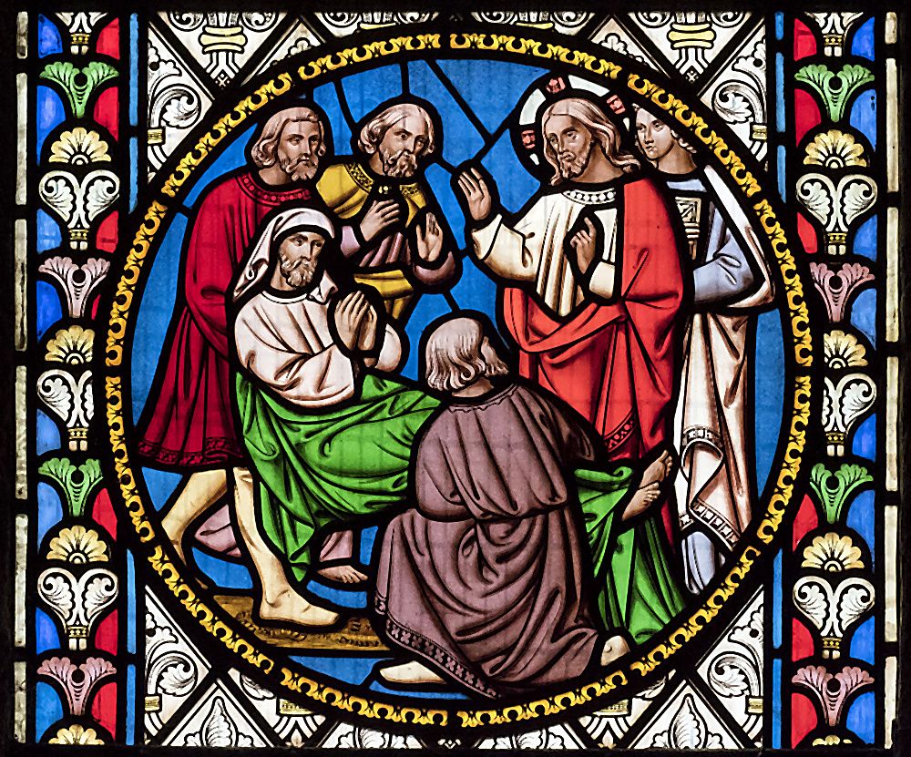 A Victorian stained-glass roundel at St Oswald's depicting a healing scene — Christ raising his hand in blessing over a kneeling figure in white robes, witnessed by apostles, against a brilliant blue background framed by Gothic foliage