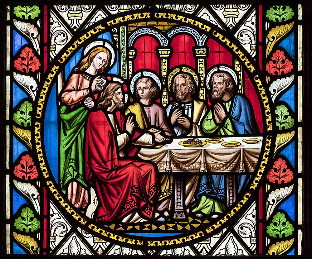 A Victorian stained-glass roundel at St Oswald's depicting the Supper at Emmaus — Christ in red and green robes seated at a draped table with the disciples, raising the bread in blessing, framed by ornate Gothic foliage and golden architectural detail