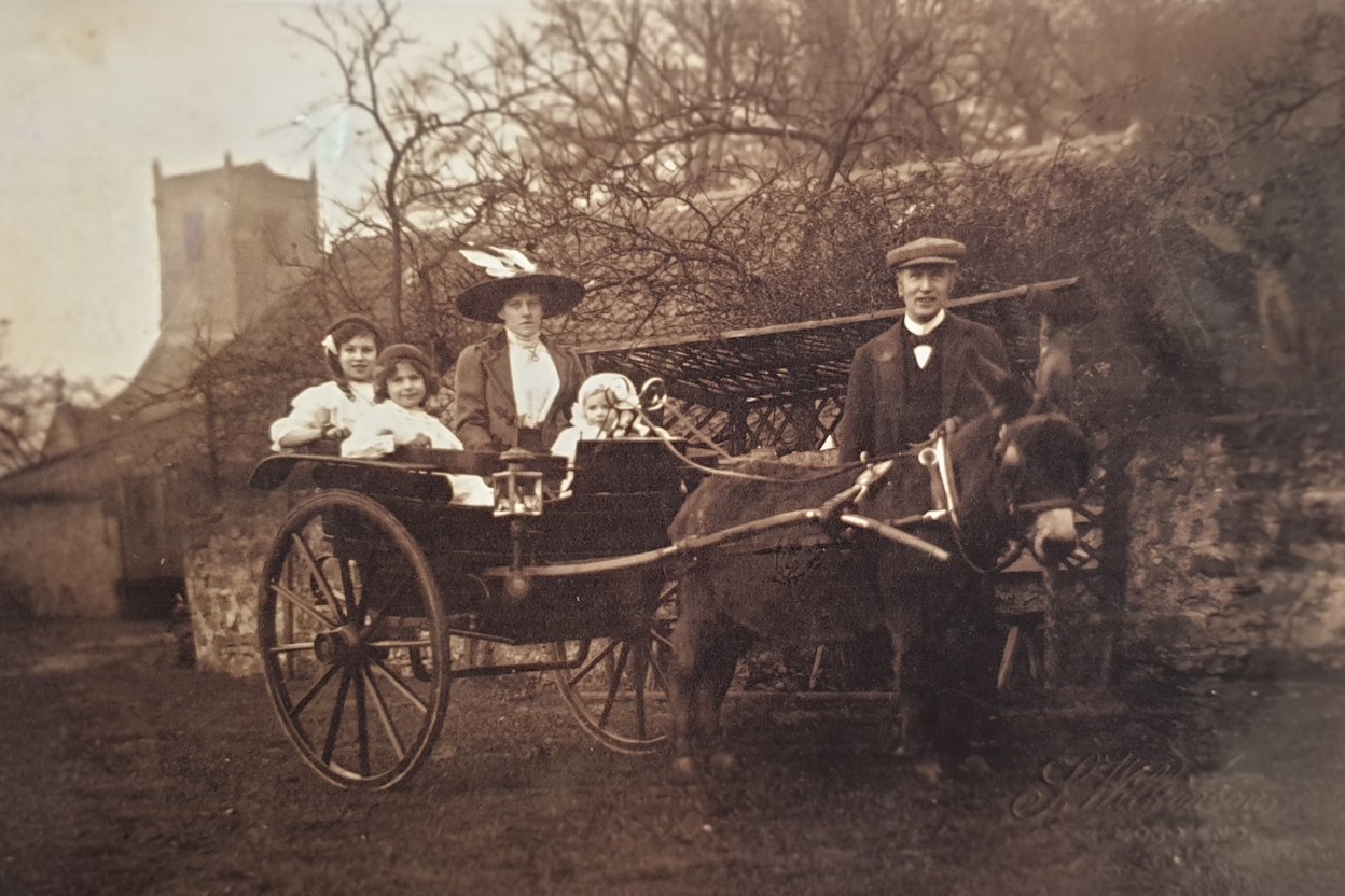 Original sepia photograph of an Edwardian family of five in a polished donkey trap. The mother sits in the trap in a wide black hat and coat, two little girls in white pinafores beside her, a baby between them; the father stands holding the donkey's head, in cloth cap and waistcoat. The fifteenth-century tower of St Oswald's rises through bare winter branches behind.