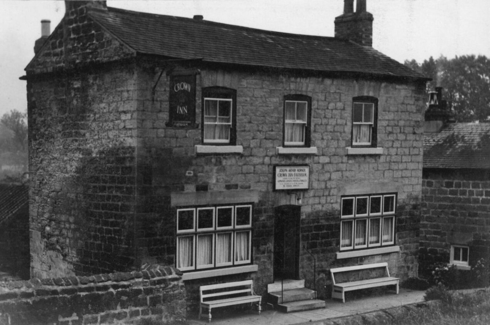 Original black-and-white photograph of the Crown Inn, Farnham, c. 1940s. A two-storey stone building with twin chimneys, the hanging 'Crown Inn' sign on the front, and two wooden benches beneath the ground-floor windows. The licensee's plaque above the door reads 'Joseph Arthur Horner, Crown Inn, Farnham'.