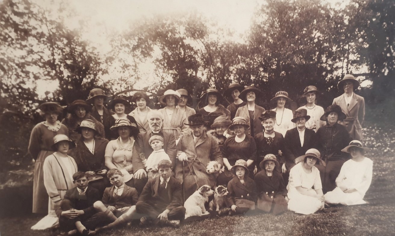Original sepia group portrait of about thirty Farnham villagers gathered on a lawn beneath summer trees. Three rows: women standing at the back in cloche hats, a middle row seated with two older bearded gentlemen in tweed and a small boy holding a posy; the children seated at the front with two small terriers. All look at the camera in formal Edwardian-into-Twenties dress.