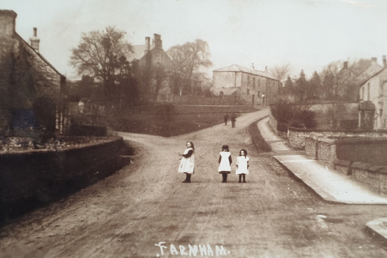Original sepia photograph of three small girls in white pinafores standing in the dust of a Farnham village lane, looking back at the camera. Stone cottages line the left side; a long farm range with rust-red roof sits to the right above stone walls. The word 'FARNHAM' is written by hand at the bottom of the photograph.