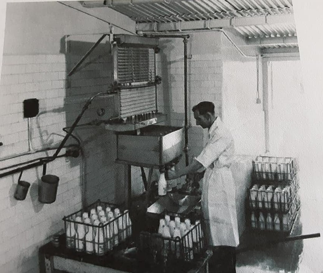 Original black-and-white photograph of the bottling room at Manor Farm Dairy. A white-coated dairyman stands at the bottling machine, feeding bottles in under the manifold. White-tiled walls, corrugated metal ceiling, full crates of bottled milk stacked along the back wall.