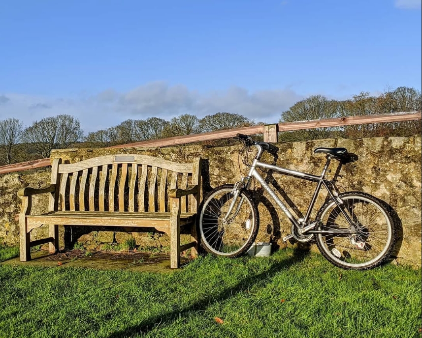A wooden memorial bench and a silver mountain bike leaning against a low coursed-stone wall, with bare winter trees in the background, on the edge of Farnham village