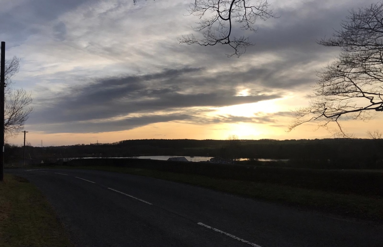 A country lane curving past the Farnham lakes at dusk, the calm water and far hills lit by a dramatic golden-orange sunset breaking through layered cloud, bare winter trees framing the view either side