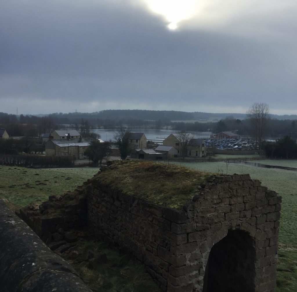 The ruined stone arch on the edge of Farnham village in foreground, with a moody overcast winter sky, looking across the village rooftops to the lake and the line of moored sailing dinghies at the Ripon Sailing Club marina