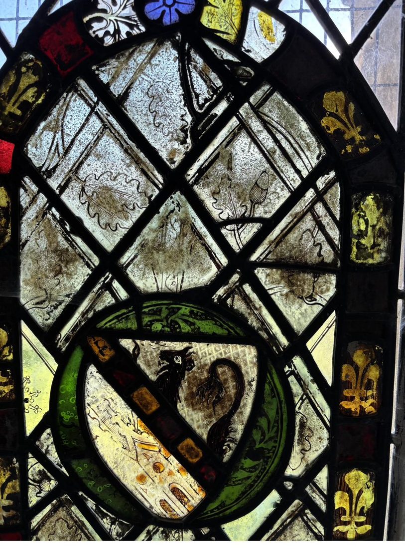 Close detail of the fourteenth-century stained glass panel at St Oswald's Church, Farnham, before restoration, showing a black-painted heraldic lion set within a green roundel, surrounded by oak-leaf quarries and a silver-stained fleur-de-lis border